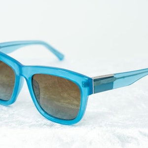 K&ouml;nnte beinhalten: Eine blaue Sonnenbrille mit braunen Gl&auml;sern. Die Sonnenbrille hat einen quadratischen Rahmen und einen silbernen Akzent auf der Br&uuml;cke.