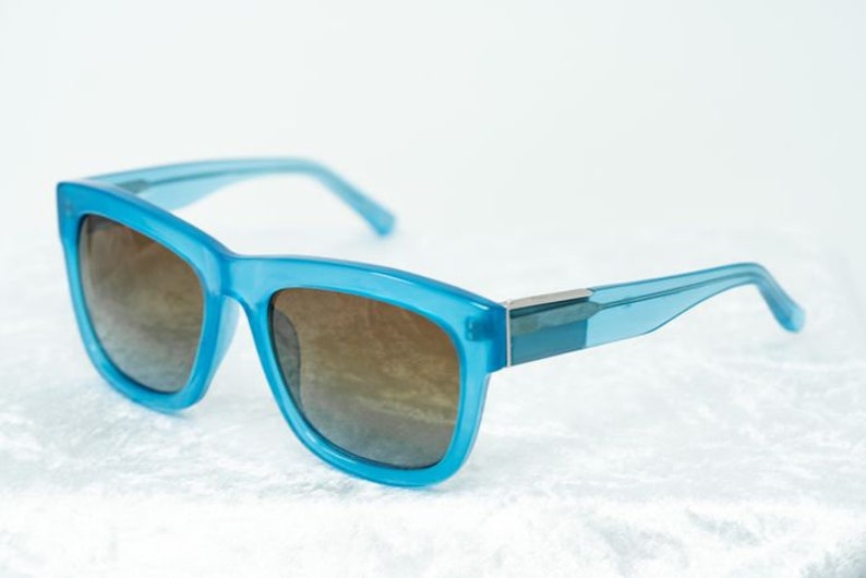 K&ouml;nnte beinhalten: Eine blaue Sonnenbrille mit braunen Gl&auml;sern. Die Sonnenbrille hat einen quadratischen Rahmen und einen silbernen Akzent auf der Br&uuml;cke.