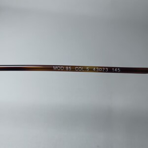 May include: Close-up of the arm of a pair of eyeglasses. The arm is a brown tortoiseshell pattern. White text on the arm reads "MOD.85 COL.5 43023 145".