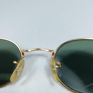 Puede incluir: Gafas de sol redondas Ray-Ban doradas con lentes verdes. Las gafas tienen un marco de metal y las palabras "62L RAY-BAN USA" est&aacute;n grabadas en el puente.