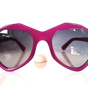 Könnte beinhalten: Hellrosa Sonnenbrille mit einem einzigartigen geometrischen Rahmen und Farbverlaufsgläsern. Die Sonnenbrille hat ein kühnes, eckiges Design und bietet einen modernen und stilvollen Look. Die Gläser gehen von dunkel nach hell über.