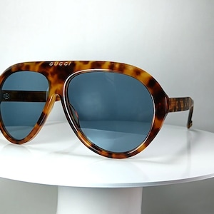 May include: Oversized aviator sunglasses featuring a brown and black tortoiseshell pattern frame with a thick top bar. The lenses are a solid dark blue tint. The word GUCCI is printed in gold lettering on the center of the top bridge of the frame.