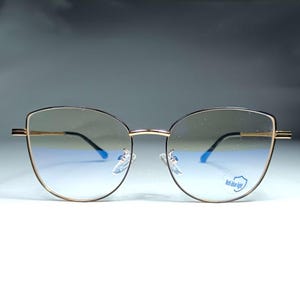 May include: Gold-rimmed cat-eye glasses with blue light blocking lenses. The lenses are tinted a light brown color. The glasses have a black frame and the words "Anti-blue light" are printed on the right lens.