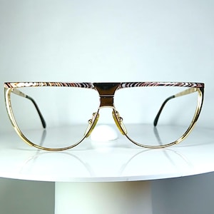 May include: Gold-toned eyeglasses with a unique, half-rim design. The frames feature a decorative pattern on the top edge and a bridge. The lenses are clear, and the arms are thin and black. The glasses are displayed on a white surface.