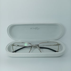 Puede incluir: Gafas de montura plateada con patillas negras en una caja blanca con la palabra "Oakley" impresa en el interior.