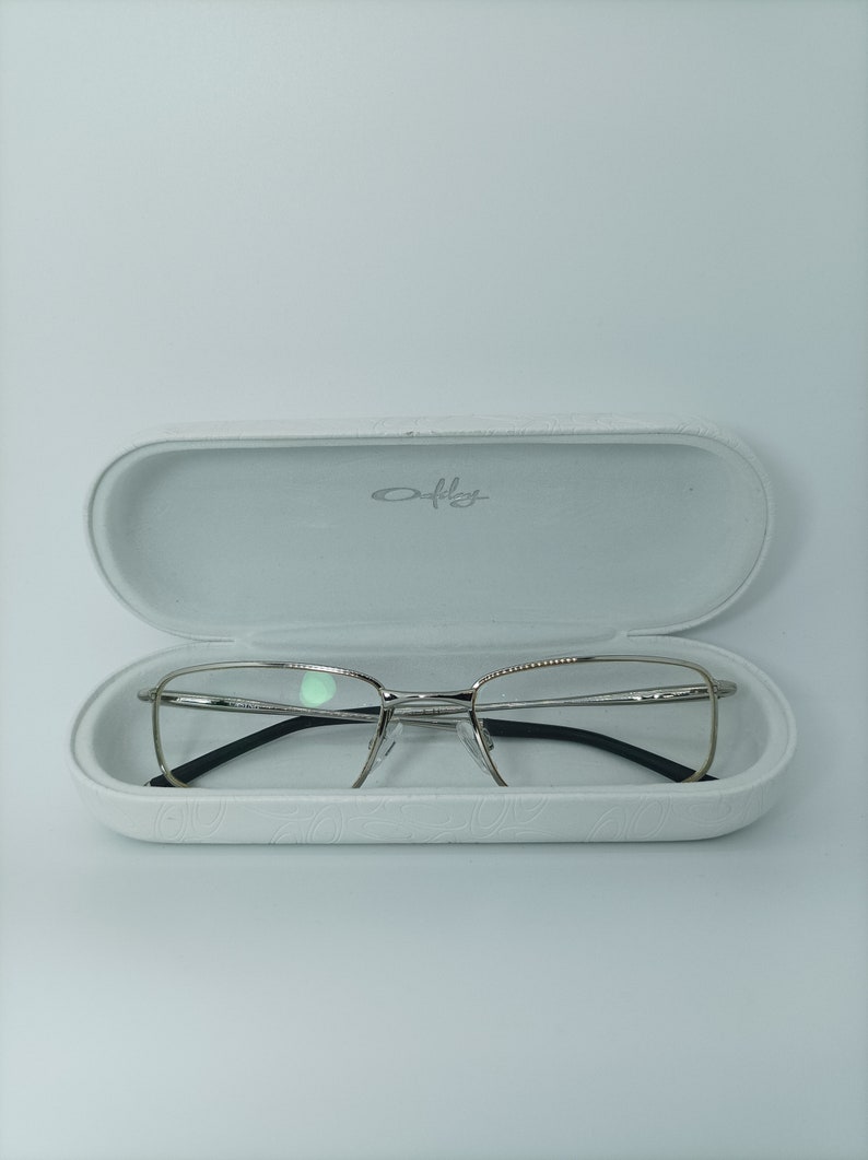 Puede incluir: Gafas de montura plateada con patillas negras en una caja blanca con la palabra "Oakley" impresa en el interior.