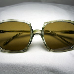 Może przedstawiać: Okulary przeciwsłoneczne vintage z przezroczystą, jasnozieloną oprawką i brązowymi szkłami. Oprawka ma kwadratowy kształt ze zaokrąglonymi rogami.