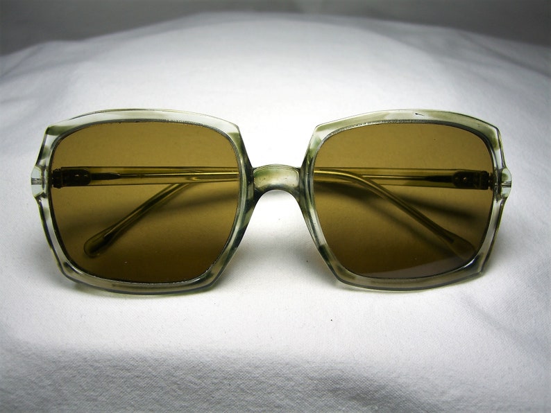 Może przedstawiać: Okulary przeciwsłoneczne vintage z przezroczystą, jasnozieloną oprawką i brązowymi szkłami. Oprawka ma kwadratowy kształt ze zaokrąglonymi rogami.