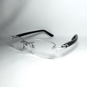 Könnte beinhalten: Randlose Brille mit klaren Gläsern und silberfarbenen Metallakzenten. Die Bügel sind schwarz und am Ohr gebogen. Die Brille ist vor einem weißen Hintergrund platziert.