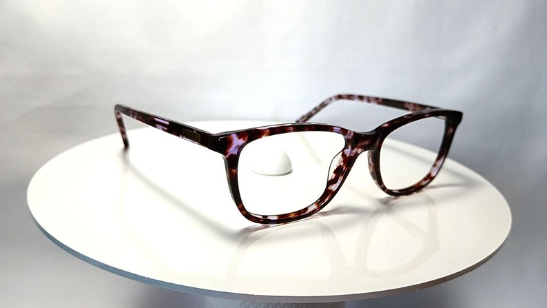 Peut inclure: Lunettes &agrave; motif &eacute;caille de tortue dans des tons de marron, noir et violet. Les montures sont de forme rectangulaire avec une l&eacute;g&egrave;re forme &oelig;il de chat. Les lunettes sont pr&eacute;sent&eacute;es sur une surface blanche.