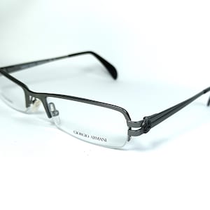 May include: Dark gray Giorgio Armani eyeglasses with a semi-rimless design. The frames have a rectangular shape with a curved lower edge. The temples are thin and feature a decorative emblem near the hinges. The lenses are clear, and the brand name is printed on the lens.