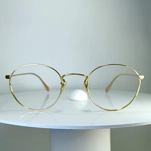 May include: A pair of gold-toned metal eyeglasses featuring thin, rounded frames and a delicate bridge. The glasses have clear lenses and slender temples that curve slightly at the ends, presenting a simple and lightweight design structure.