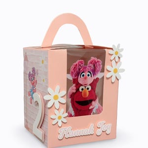 May include: A pink birthday gift box featuring Elmo and Abby Cadabby plush toys. The box has a handle, a clear window, and is decorated with daisy cutouts and the name "Hannah Joy". A large white number "2" is on the side.