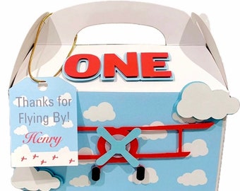 May include: A blue and white favor box with a red airplane and clouds. The box has a handle and a tag that says "Thanks for Flying By! Henry". The box also has the word "ONE" in red letters.