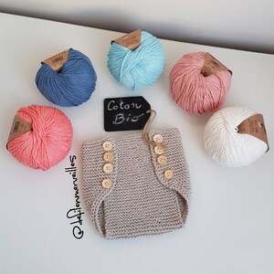 Peut inclure: Un lange bébé tricoté beige avec des boutons en bois. Cinq pelotes de laine de différentes couleurs sont disposées autour du lange. La laine est étiquetée "Coton Bis".