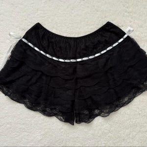 Op de afbeelding: Zwarte kanten short met meerdere gerimpelde lagen en een geschulpte zoom. Een wit lint met een decoratief stiksel detail siert de taille. Witte satijnen linten zijn aan weerszijden van de short geknoopt.