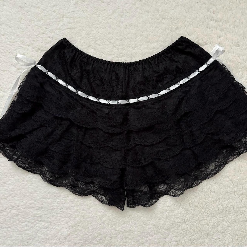Op de afbeelding: Zwarte kanten short met meerdere gerimpelde lagen en een geschulpte zoom. Een wit lint met een decoratief stiksel detail siert de taille. Witte satijnen linten zijn aan weerszijden van de short geknoopt.