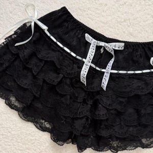 Op de afbeelding: Zwarte kanten shorts met meerdere gerimpelde lagen. De shorts hebben witte lintaccenten en witte strikken met muzieknoten erop gedrukt. De shorts zijn gemaakt van een delicaat, doorschijnend kant.