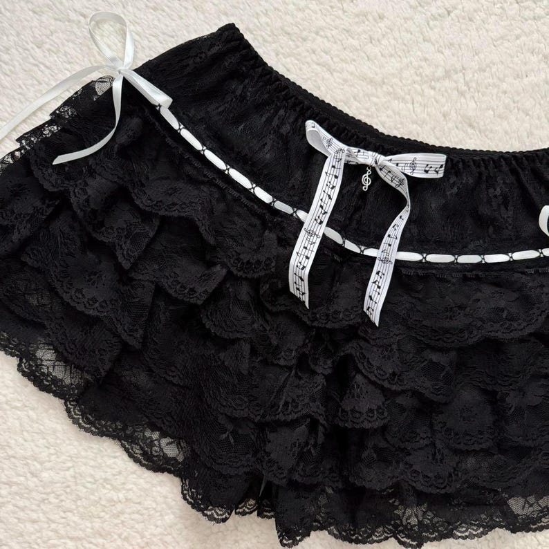 Op de afbeelding: Zwarte kanten shorts met meerdere gerimpelde lagen. De shorts hebben witte lintaccenten en witte strikken met muzieknoten erop gedrukt. De shorts zijn gemaakt van een delicaat, doorschijnend kant.