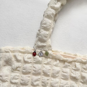 May include: Close-up of a cream-colored textured top with a decorative pin. The pin features a red ladybug charm, a white circle with the letter "L", and a green leaf charm. The top has a square pattern.