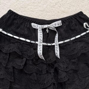 Op de afbeelding: Zwarte kanten rok met meerdere gerimpelde lagen. Een wit lint met muzieknoten en een G-sleutel-bedel siert de voorkant. De rok heeft een witte rand met kleine decoratieve accenten.