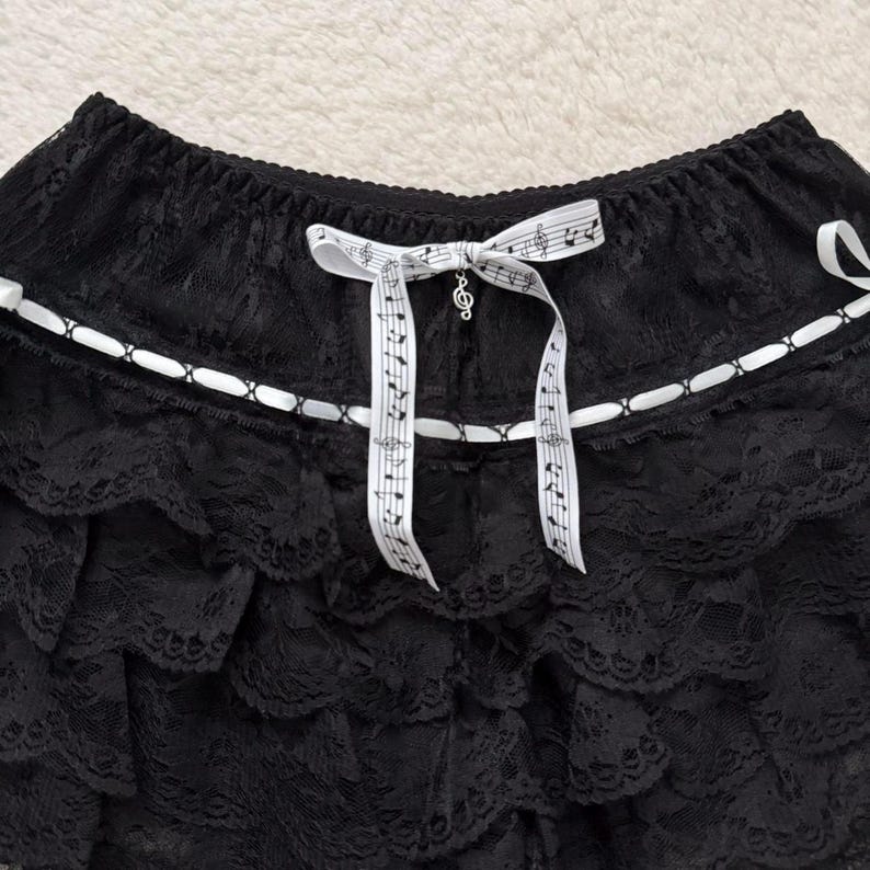 Op de afbeelding: Zwarte kanten rok met meerdere gerimpelde lagen. Een wit lint met muzieknoten en een G-sleutel-bedel siert de voorkant. De rok heeft een witte rand met kleine decoratieve accenten.