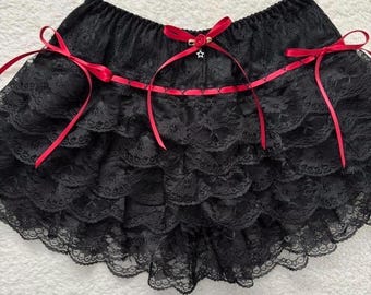 Puede incluir: Shorts de encaje negro con múltiples capas con volantes. Cintas de raso rojo atadas con lazos en la cintura y a lo largo del volante superior. Una pequeña rosa roja y un charm de estrella adornan el frente.