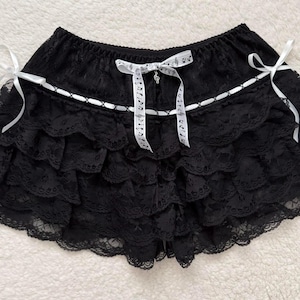 Op de afbeelding: Zwarte kanten shorts met meerdere gerimpelde lagen. De shorts hebben witte lintaccenten en strikken, waaronder een strik met muzieknoten. Een kleine bedel hangt aan de muzieknotenstrik. De shorts hebben een elastische tailleband.