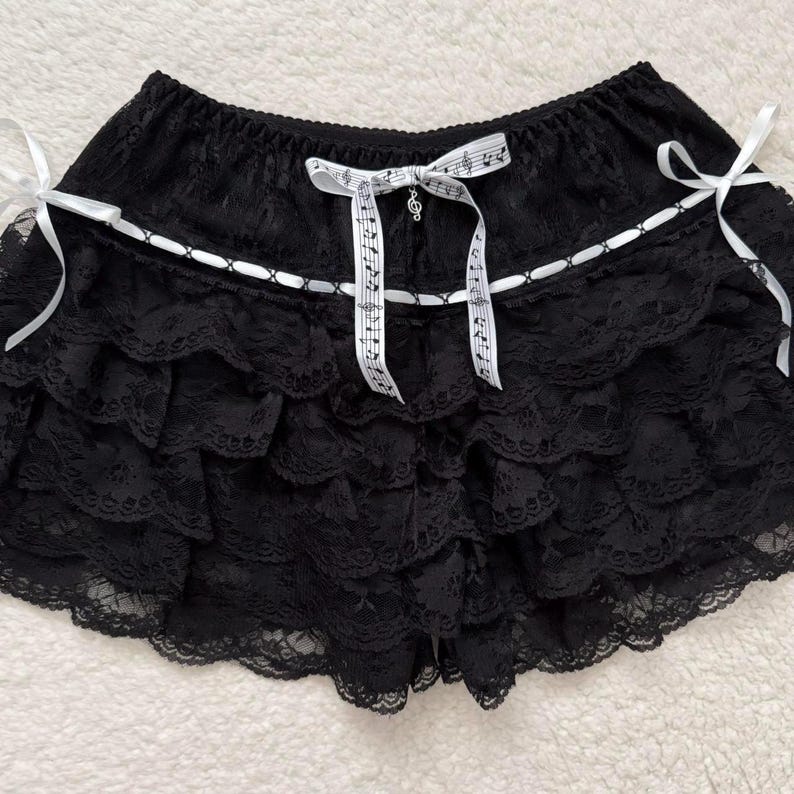 Op de afbeelding: Zwarte kanten shorts met meerdere gerimpelde lagen. De shorts hebben witte lintaccenten en strikken, waaronder een strik met muzieknoten. Een kleine bedel hangt aan de muzieknotenstrik. De shorts hebben een elastische tailleband.