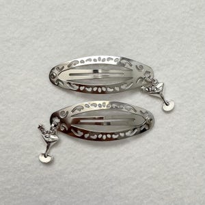 May include: Two silver-toned hair barrettes with an oval shape and intricate cut-out designs. Each barrette has a small charm of a martini glass hanging from it. The barrettes are on a white background.