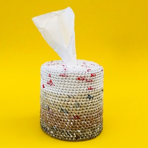 Pode incluir: Uma capa para caixa de lenços de tecido de crochê branco com um padrão marrom, bege e vermelho. A capa tem um lenço de papel branco que está saindo do topo.