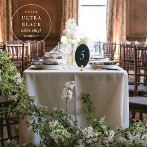 Peut inclure: Une table de r&eacute;ception avec une nappe blanche, des assiettes &agrave; motifs noirs et blancs et un num&eacute;ro de table noir avec le chiffre 5 en blanc. La table est d&eacute;cor&eacute;e de fleurs blanches et de verdure. Le texte de l'image indique "PAPER ULTRA BLACK white vinyl number."