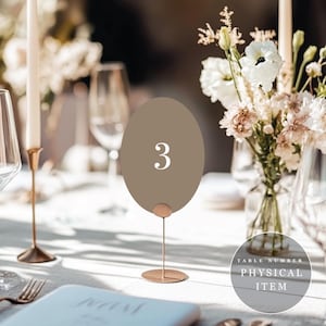 May include: An oval-shaped taupe table number sign featuring the white numeral 3, supported by a thin metallic rose gold stand with a circular base. The sign is positioned upright on a table, serving as a functional piece for event seating.