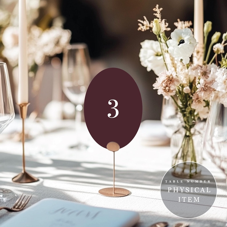 Peut inclure: Un num&eacute;ro de table ovale bordeaux avec le chiffre 3 en blanc. Le num&eacute;ro est maintenu par un fin support en m&eacute;tal dor&eacute;. L'arri&egrave;re-plan comprend une table dress&eacute;e avec des fleurs, des bougies et de la verrerie, sugg&eacute;rant un mariage ou un &eacute;v&eacute;nement.