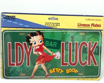 Peut inclure: Plaque d'immatriculation Betty Boop avec les mots "LDY LUCK". La plaque a un fond vert avec des dés et d'autres graphiques. Betty Boop porte une robe rouge et tient des cartes à jouer. L'emballage indique "Collector Series License Plates".