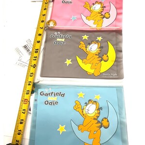 May include: Three clear plastic pouches featuring Garfield and Odie. Each pouch has a different background color: pink, gray, and blue. Garfield is depicted sitting on a crescent moon with stars. The text "Garfield and Odie" and "Starry Night" are visible.