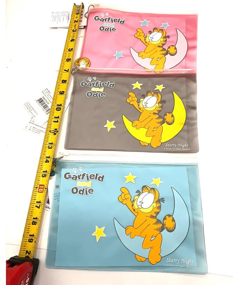 May include: Three clear plastic pouches featuring Garfield and Odie. Each pouch has a different background color: pink, gray, and blue. Garfield is depicted sitting on a crescent moon with stars. The text "Garfield and Odie" and "Starry Night" are visible.