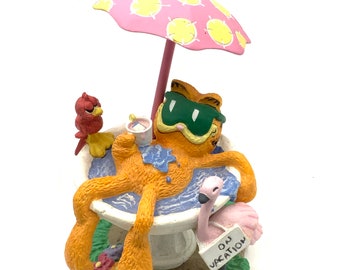 May include: A figurine of Garfield the cat relaxing in a bird bath with a pink flamingo, a red bird, and a pink umbrella. The sign says "On Vacation".