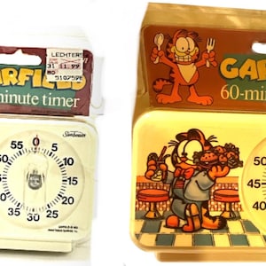 May include: Two Sunbeam Garfield 60-minute kitchen timers in original packaging. The left timer is cream-colored with Garfield and the text "TIMES UP!! LET'S EAT!". The right timer is yellow with Garfield holding a tray of food in a diner setting.