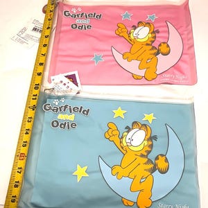 May include: Two clear plastic pouches featuring Garfield and Odie. One pouch is pink with blue stars, the other is blue with yellow stars. Both pouches have Garfield sitting on a crescent moon with the words "Starry Night".