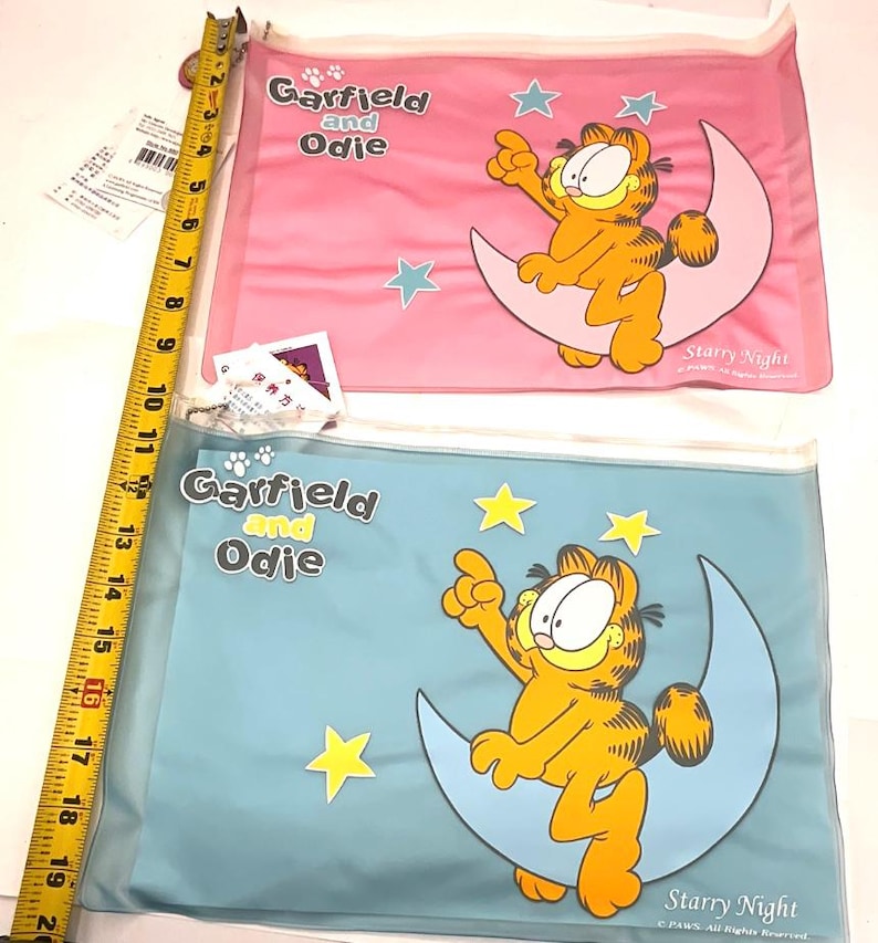 May include: Two clear plastic pouches featuring Garfield and Odie. One pouch is pink with blue stars, the other is blue with yellow stars. Both pouches have Garfield sitting on a crescent moon with the words "Starry Night".