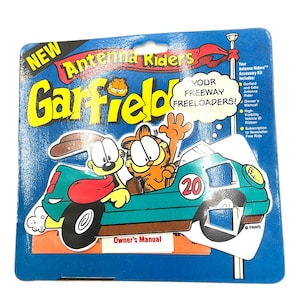 May include: A blue and orange cardboard box with a Garfield Antenna Riders toy inside. The toy features Garfield and Odie driving a green race car with the number 20 on the side. The box includes the text "New Antenna Riders Garfield Your Freeway Freeloaders!" and "Owner's Manual".