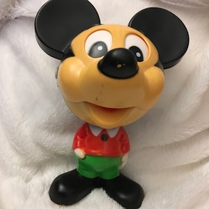May include: A vintage plastic Mickey Mouse toy with black ears, a yellow face, red shirt, and green pants. The toy has a smiling expression and large eyes.
