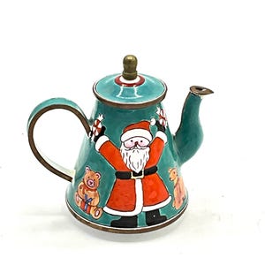 May include: A teal blue enamel teapot with a brass lid and handle. The teapot is decorated with a Christmas scene featuring Santa Claus, a teddy bear, and a reindeer.