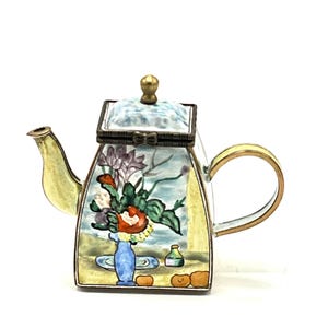 May include: A small, square, yellow teapot with a floral design and a hinged lid. The teapot has a spout and a handle. The floral design includes a blue vase with flowers and oranges.