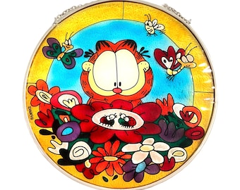 May include: Round stained glass window featuring Garfield the cat surrounded by flowers and butterflies. The window has a yellow border, a blue background, and a silver chain for hanging. The artwork is colorful and whimsical.