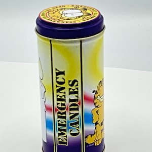 May include: A yellow and purple cylindrical tin with a cartoon cat on the front. The tin has the words "Emergency Candles" printed on the side. The lid of the tin has a cartoon cat on it.
