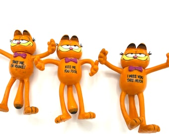 Può includere: Tre figurine di gatto Garfield arancione con braccia e gambe flessibili. Ogni figurina ha un messaggio diverso sul petto: "Prendimi, sono tuo!" , "Baciami, stupido", e "Mi manchi tanto".