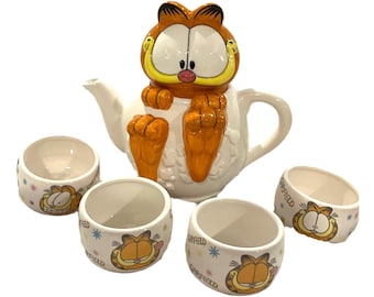 May include: A white ceramic Garfield-themed tea set. The teapot features Garfield's face and body. The set includes four matching cups with Garfield's face and the word "Garfield".