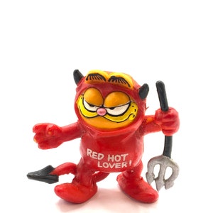 May include: Red devil Garfield figurine with a pitchfork. He is wearing a red devil costume with a tail and horns. The costume has the words "Red Hot Lover!" printed on the front.